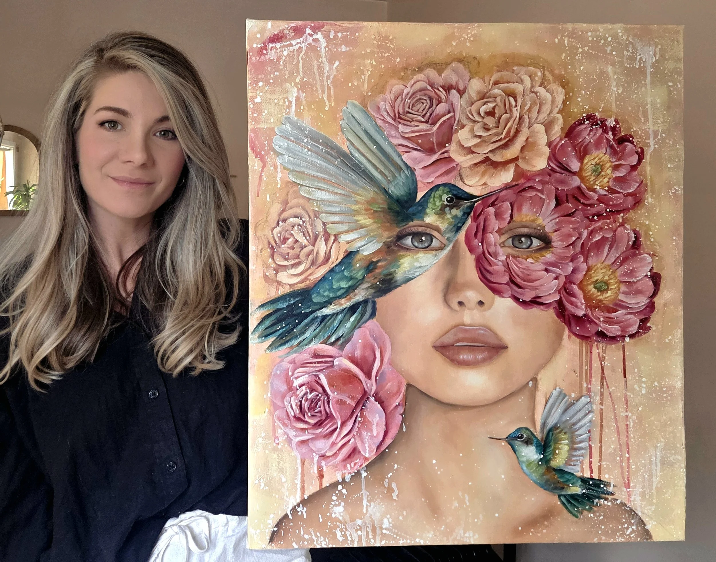 A woman with long blonde hair taking a selfie beside a colorful painting of a woman's face, with flowers and hummingbirds, on a pink and gold background.