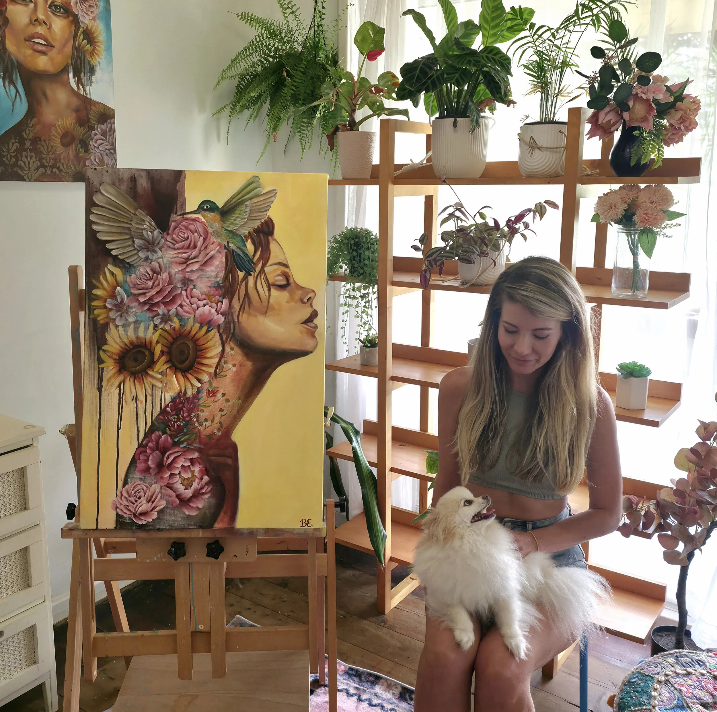 A woman with long blonde hair holding a fluffy dog in her lap next to a wooden shelving unit filled with potted plants and flowers. There is a vibrant painting of a woman's face with floral elements on the wall behind her.