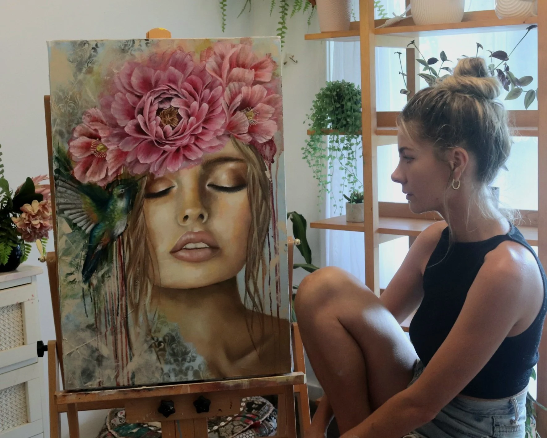 A woman sitting on the floor in front of a painting of a woman's face with eyes closed, big pink flowers on her head, and a hummingbird near her lips, in a room with wooden shelves and potted plants.