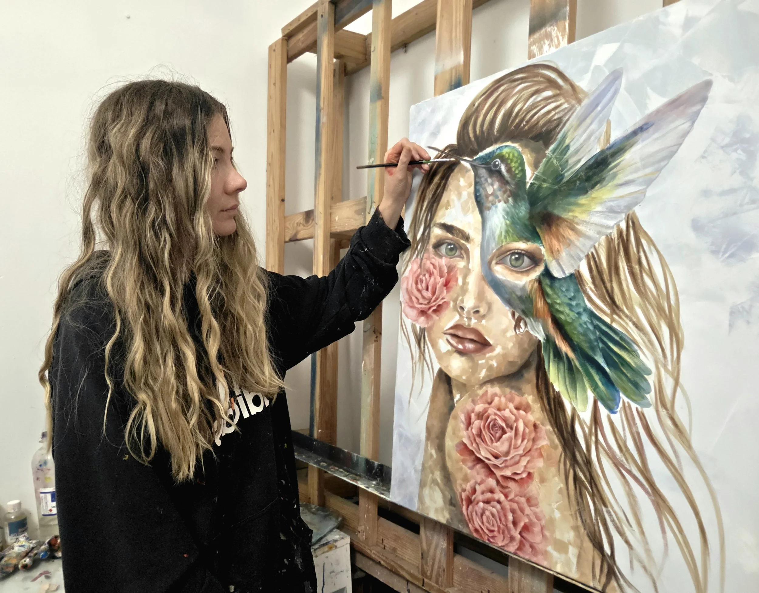 A woman with long wavy blonde hair painting a large portrait of a woman's face with transformative elements, including a colorful bird on her head and pink roses near her chin, in an art studio.