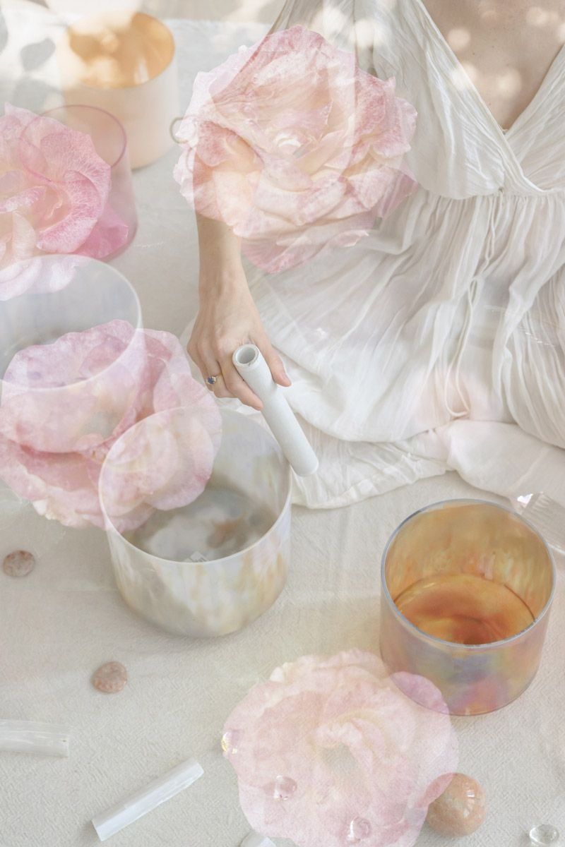 A person in a white dress is sitting on the floor, holding a white cylindrical object, surrounded by iridescent bowls and pink, peach, and white decorative paper flowers.