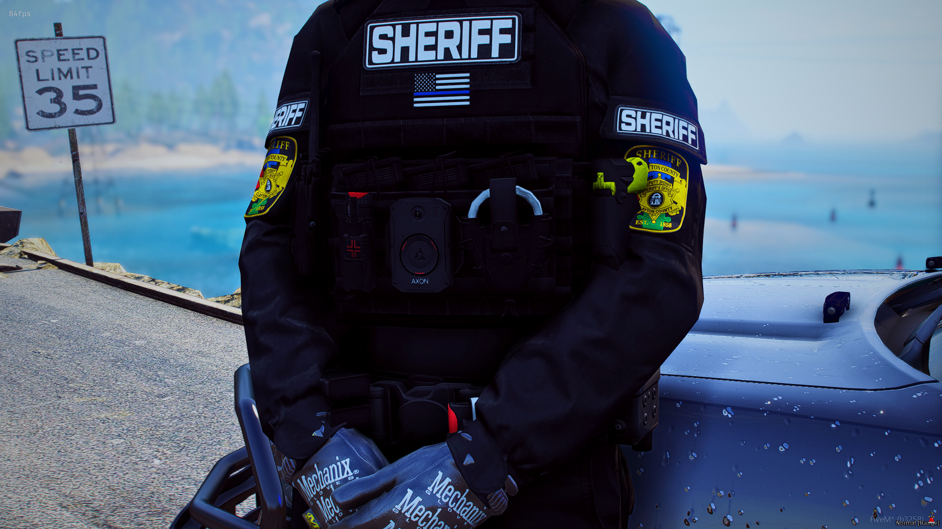 Close-up of a sheriff's uniform with patches and badges, standing next to a police vehicle, with a road sign indicating a speed limit of 35 miles per hour in the background, and water in the distance.