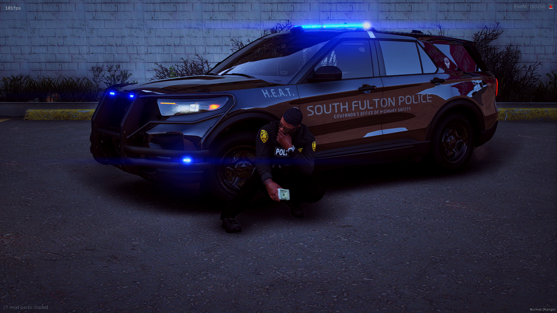 A police officer kneeling next to a police car with blue emergency lights on, holding money, parked against a brick wall with some plants behind it.