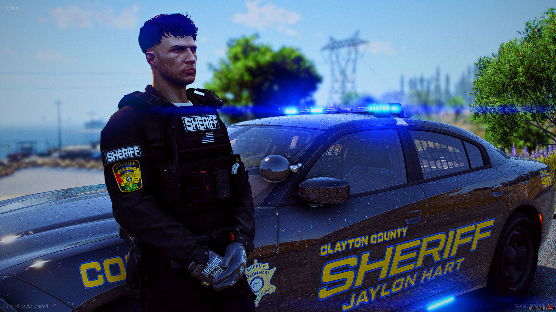 A sheriff officer standing next to a Clayton County sheriff police car with blue lights on, outdoors on a sunny day with trees and power lines in the background.