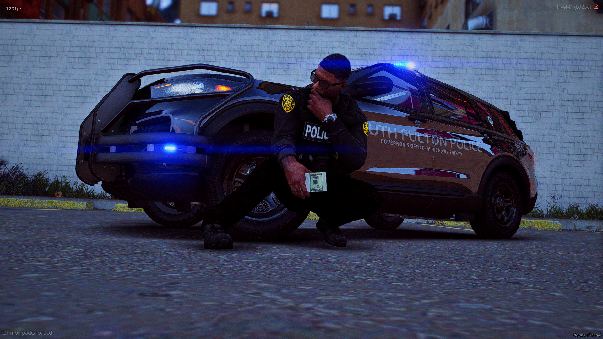 A police officer in a black uniform is kneeling on one knee beside a police SUV with its emergency lights flashing. The officer is holding a stack of U.S. $100 bills and appears to be inspecting them, with a hand on his chin. They are in an urban set
