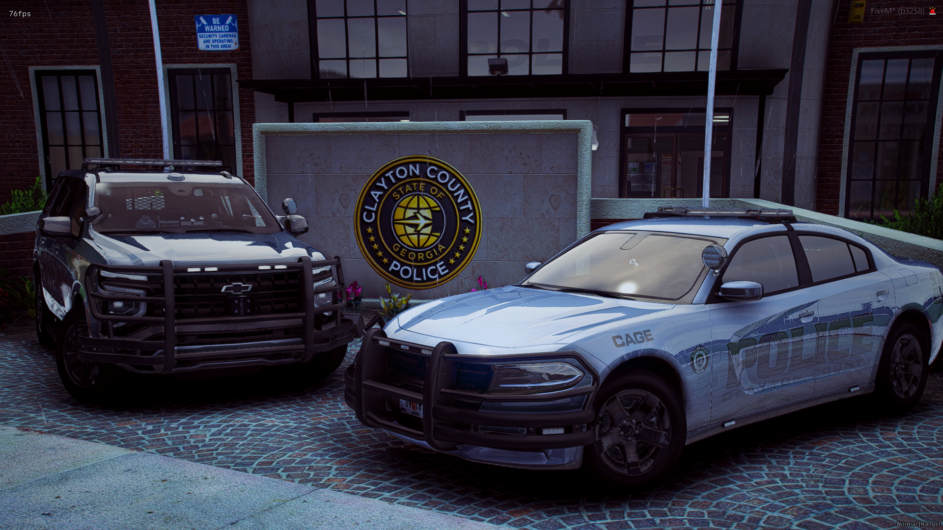 Two police cars parked outside the Clayton County Police Department building. One car is predominantly white with green markings, and the other is a black Chevrolet SUV with police decals. The building has a sign that reads "Clayton County, Georgia P