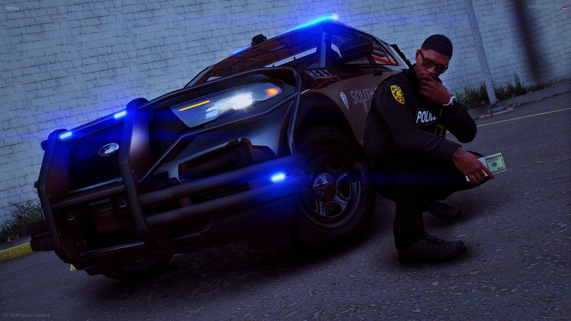 A police officer squatting by a police SUV with blue flashing lights, holding a hundred-dollar bill, in front of a brick wall.