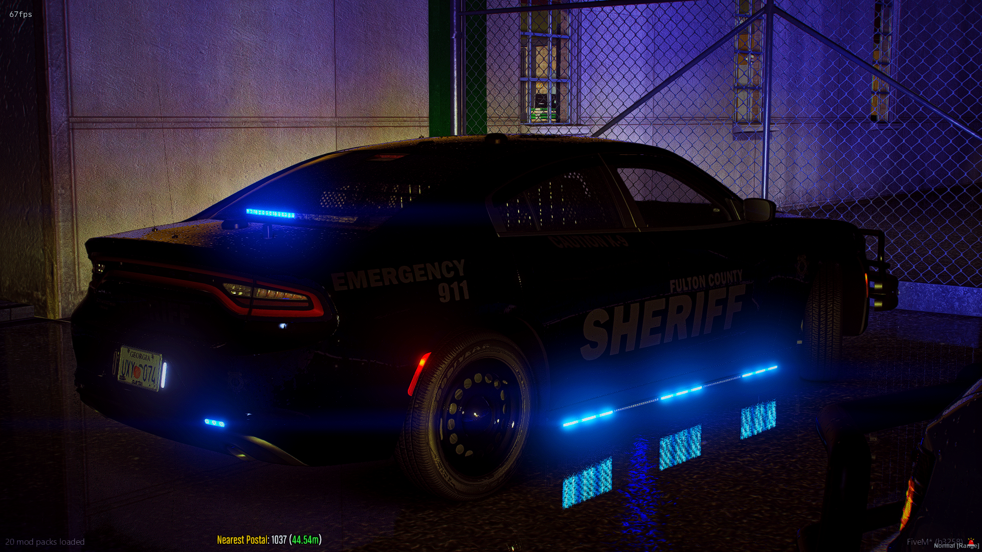 Nighttime scene of a black sheriff's patrol car with flashing blue lights parked near a chain-link fence, with water on the ground reflecting the lights.