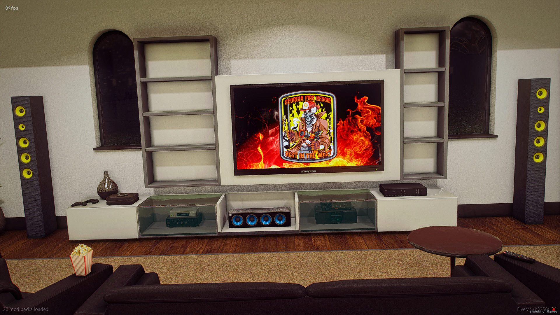 Living room with a large flat-screen TV displaying a Fire Rescue emblem, flanked by tall speaker towers, with a popcorn bucket on a black armrest sofa, a small round table, and various electronic devices.
