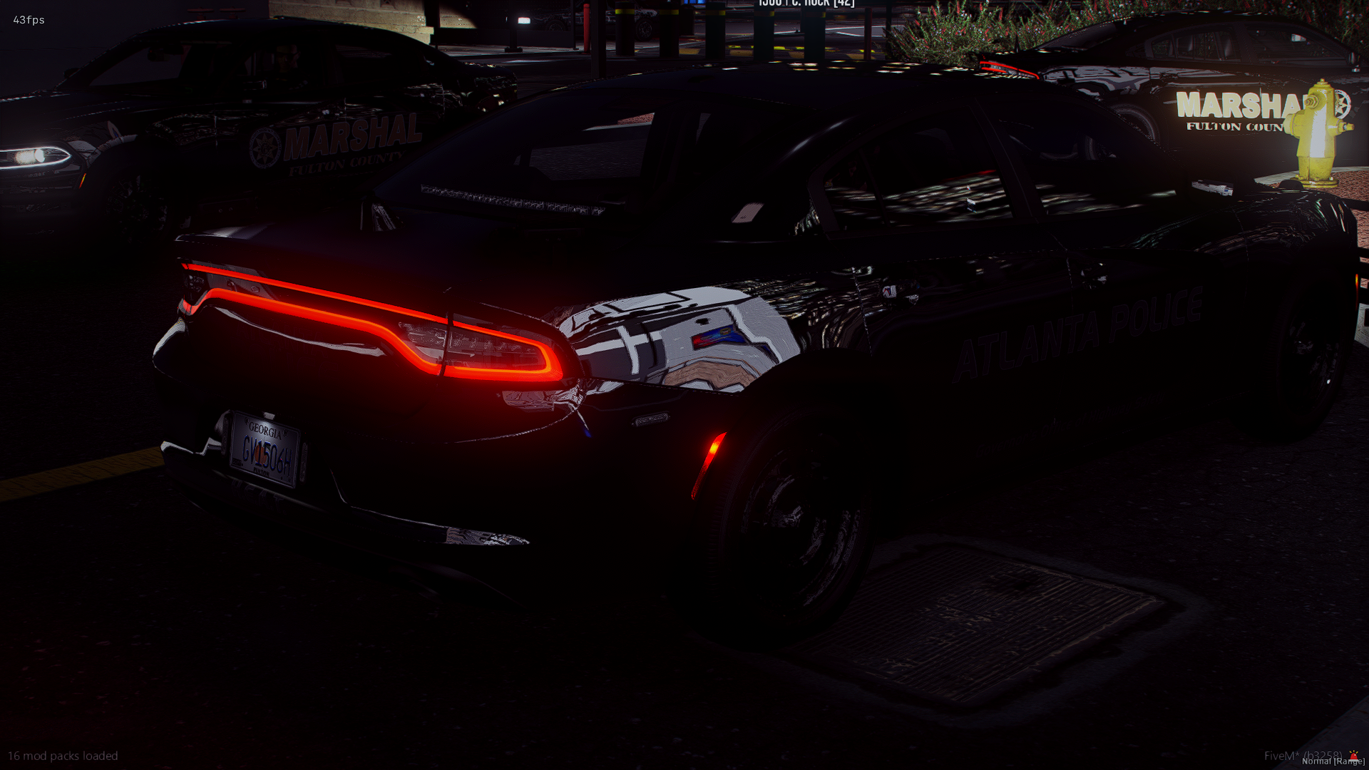 Dark-colored police car belonging to the Atlanta Police Department with illuminated tail lights, parked outdoors near other police vehicles at night, with a fire hydrant and some greenery visible in the background.