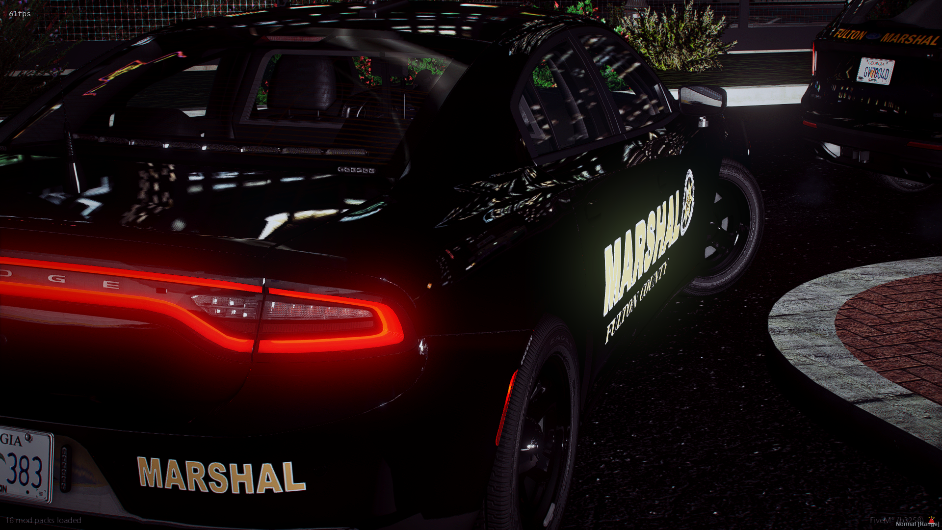 A black police car with illuminated red rear lights parked beside a curb at night. The car has the word 'MARSHAL' and a badge logo on its side, and there is a reflective pattern on the vehicle's surface. The background includes some greenery and othe