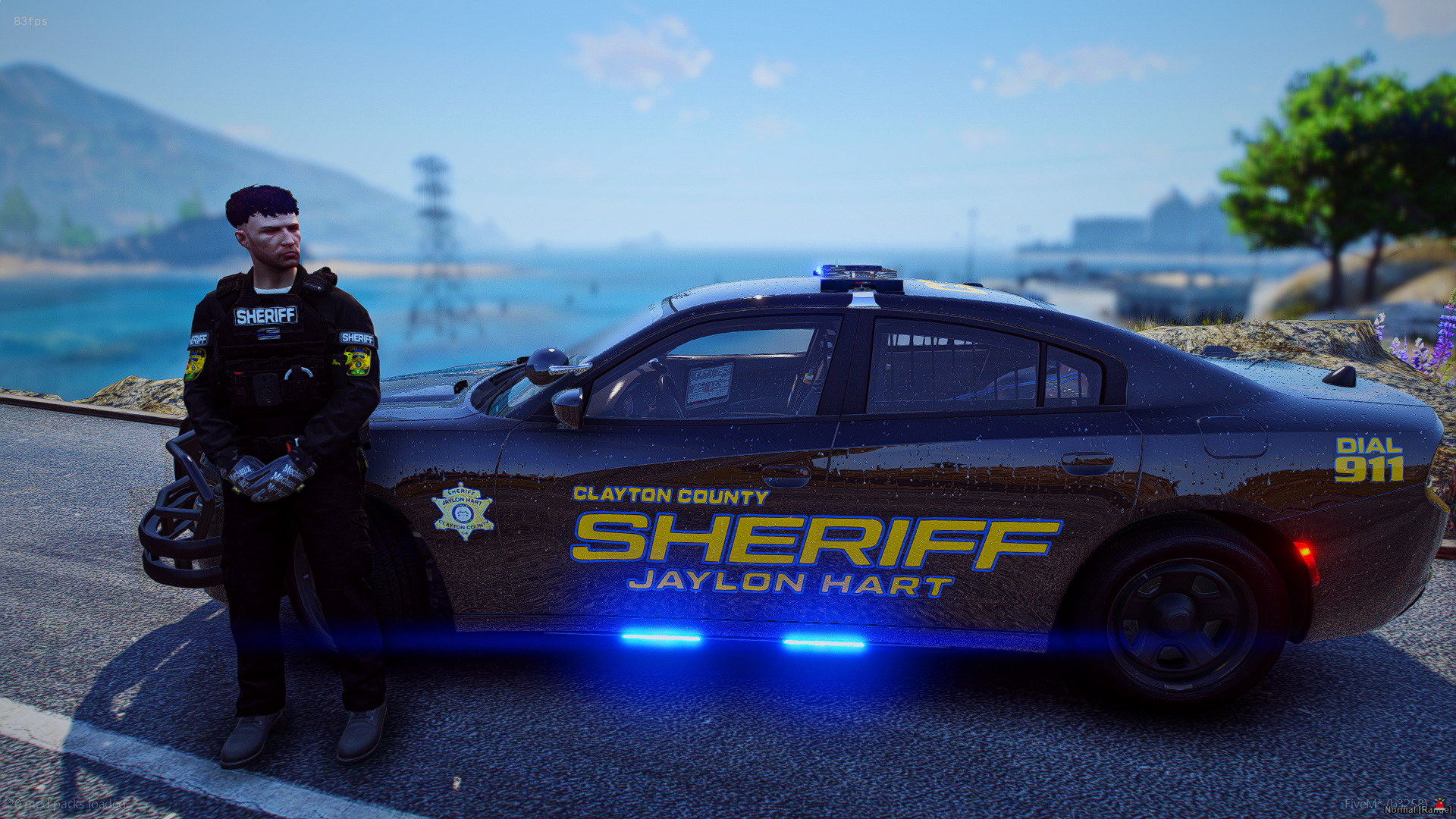 A sheriff's deputy in uniform standing beside a black Clayton County sheriff car on a road with a body of water, hills, and a tree in the background. The vehicle has blue emergency lights on.