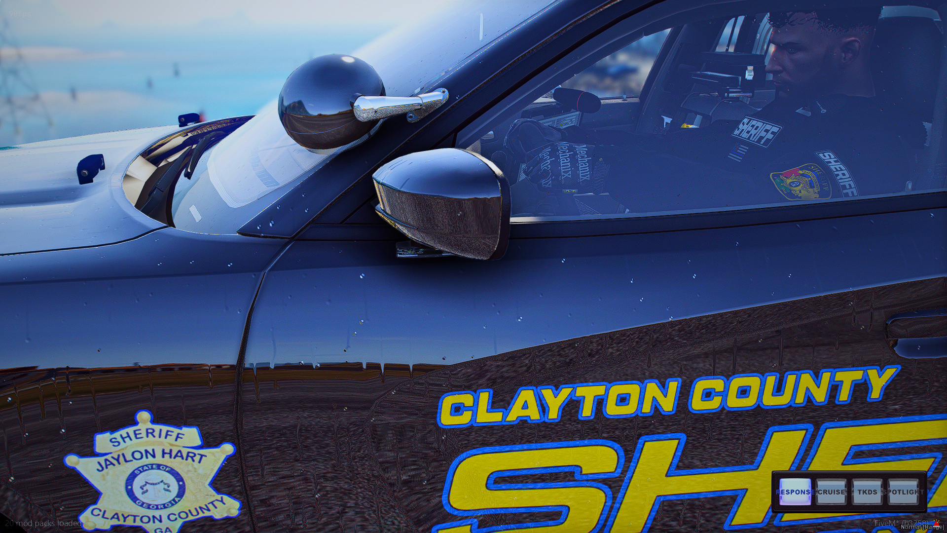 Close-up of a race car with Clayton County Sheriff's Office markings, driver inside wearing a helmet and suit, water droplets on the car, tall bridge or structure in the background.