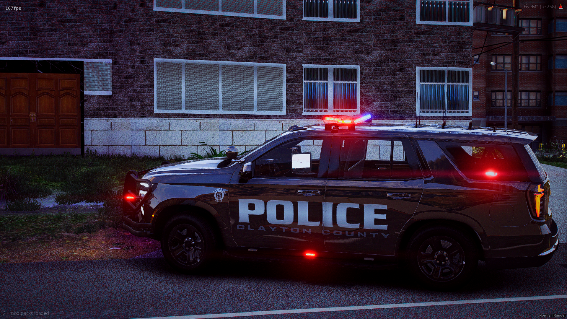 A Clayton County police SUV parked on a street at night with its red and blue emergency lights flashing. The vehicle is black with police markings, and the background shows a red brick building with several windows and a wooden door.