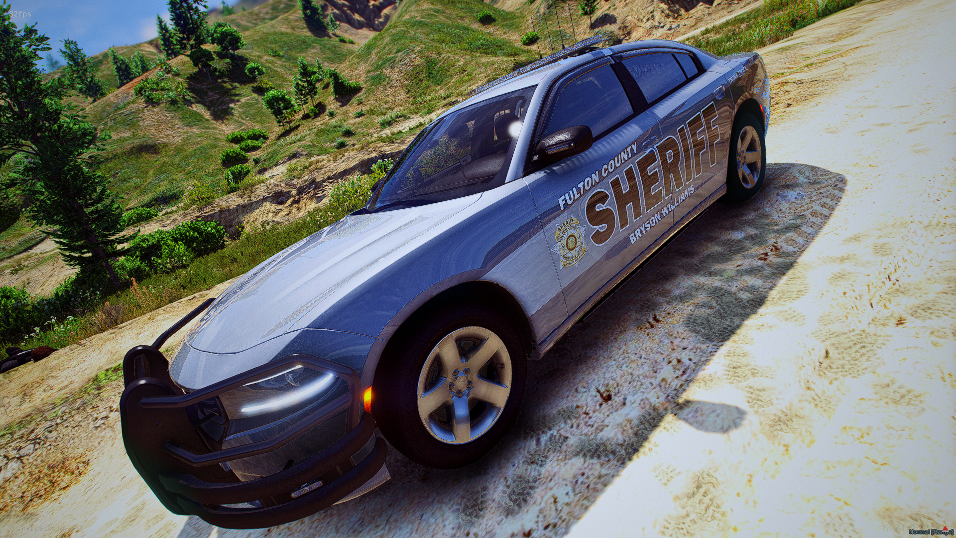 Police car labeled 'Fulton County Sheriff's' parked on a dirt road with green hills and trees in the background.