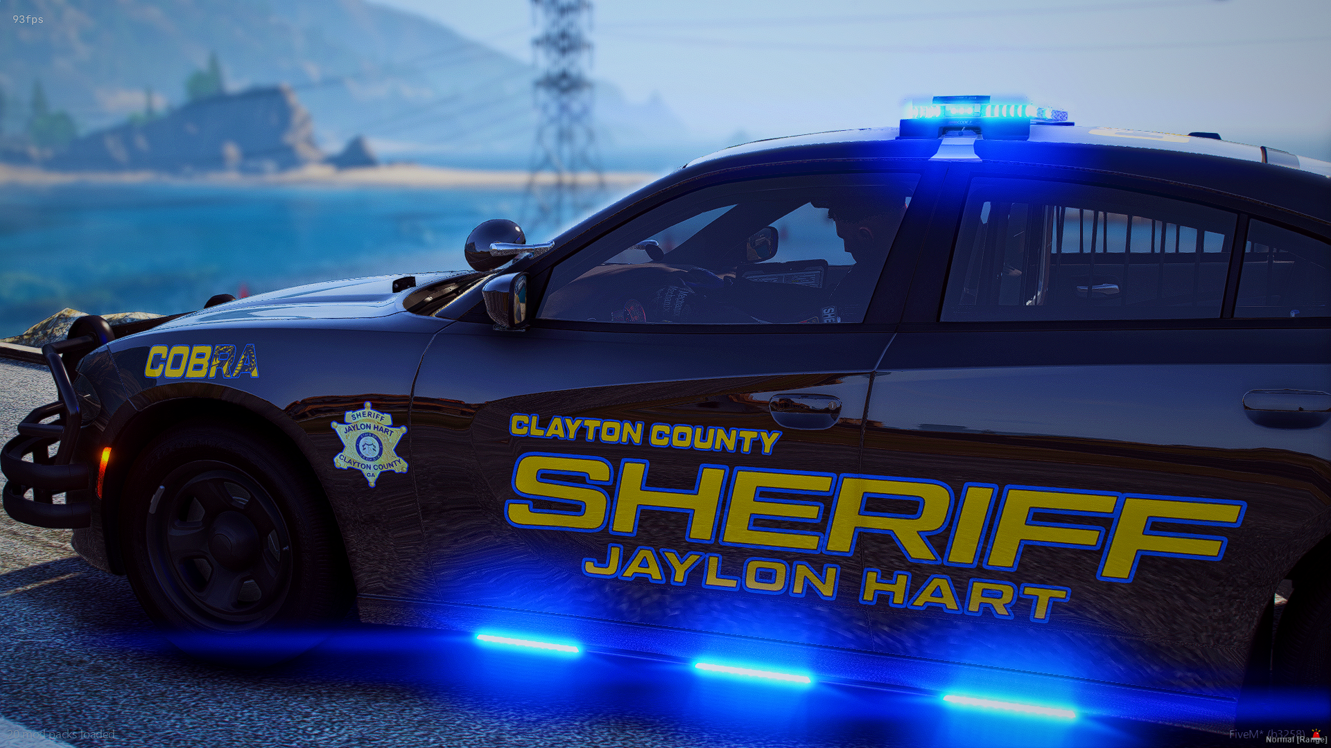 Black Clayton County Sheriffs vehicle with blue police lights on top, parked outdoors near water and power lines, with sheriff's badge and Jaylon Hart visible on the side.