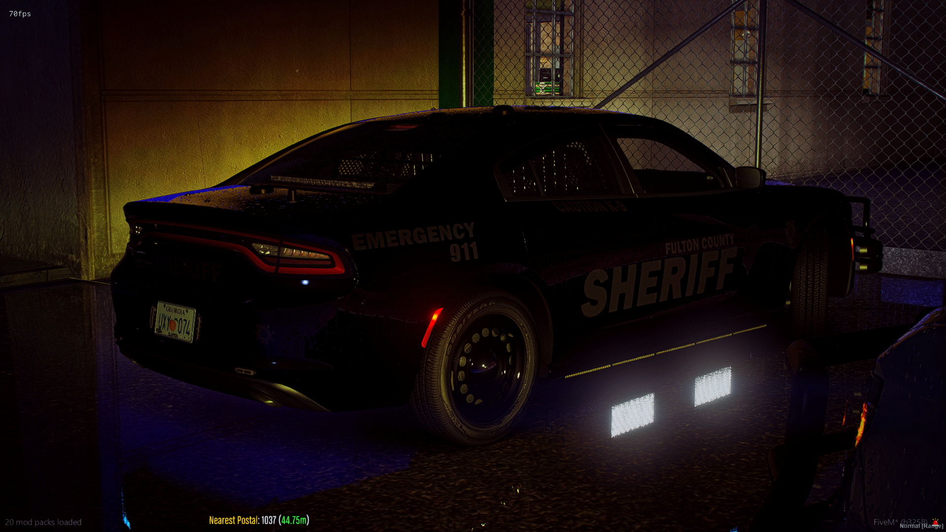 A dark-colored sheriff's patrol car parked behind a chain-link fence with a brick building in the background, illuminated by dim lights at night.