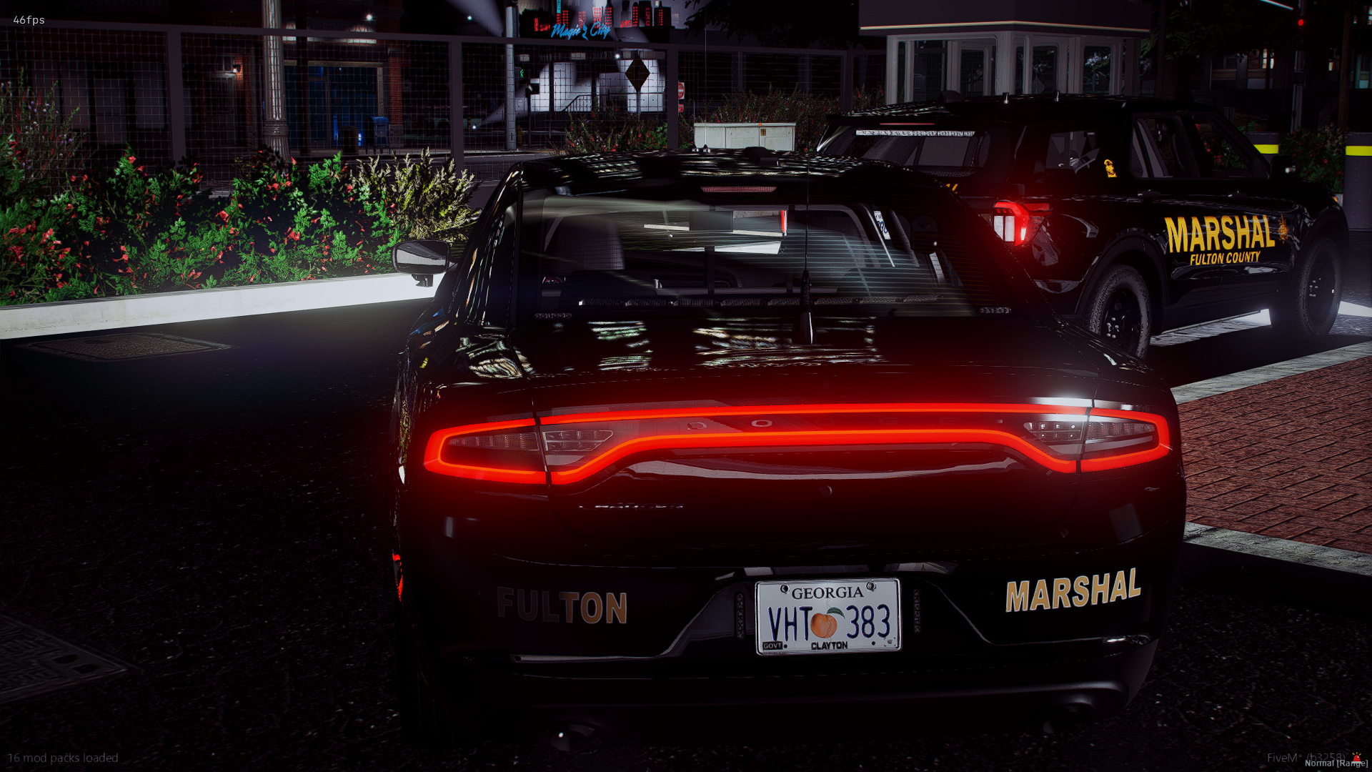 Nighttime scene showing two police SUVs with 'Fulton County Marshal' markings, parked near a landscaped area with plants and a mesh fence. The rear of a black sports car is visible in the foreground, with illuminated taillights and a Georgia license 