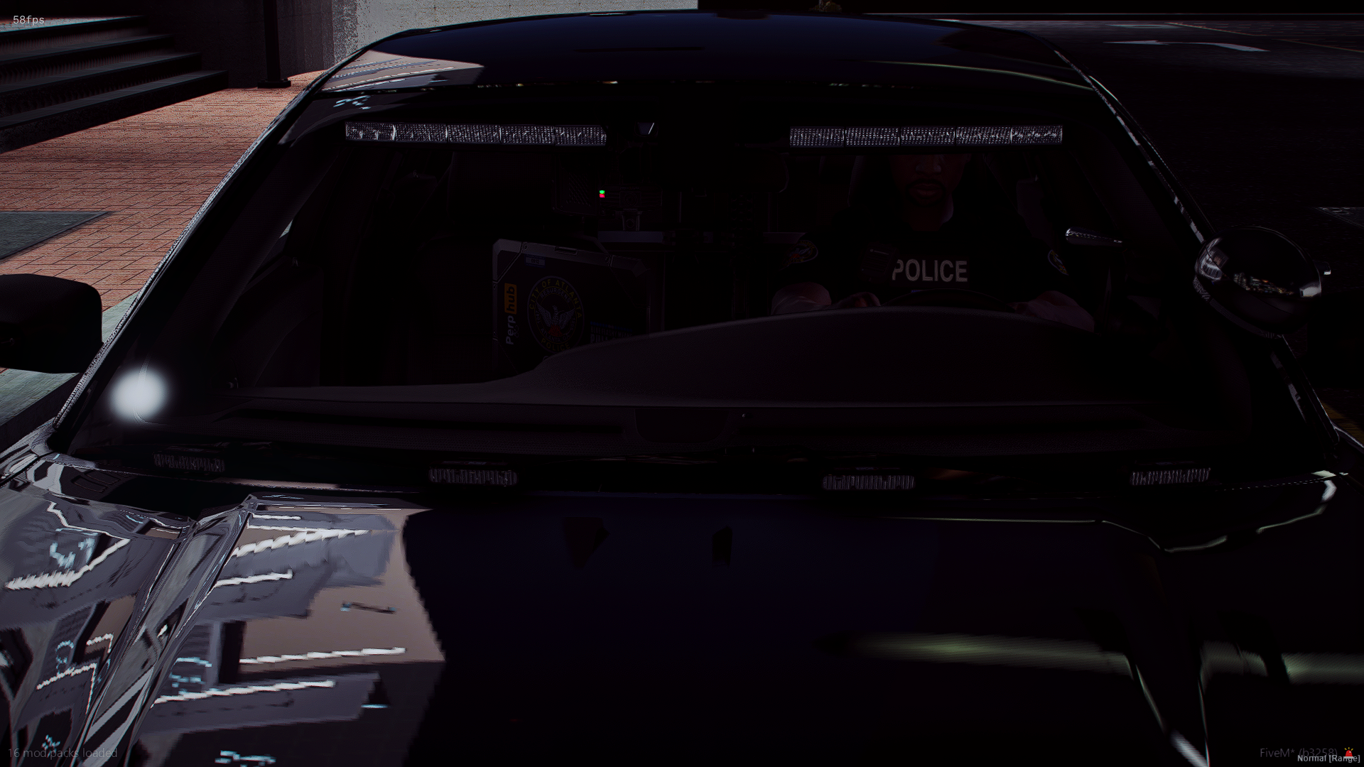 A black police car with a police officer inside, seen through the windshield. The car is parked on a brick-paved area near stairs, with part of the surrounding building visible.