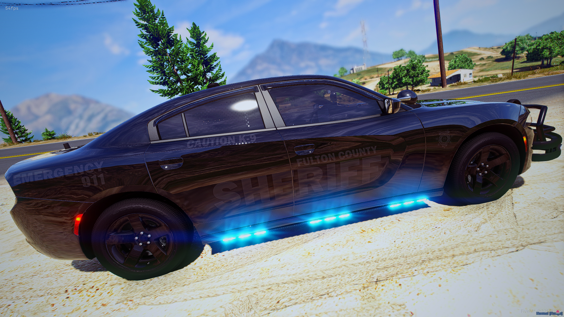 Black sheriff patrol car with blue emergency lights on the side, parked on the side of a rural road with trees, hills, and mountains in the background.
