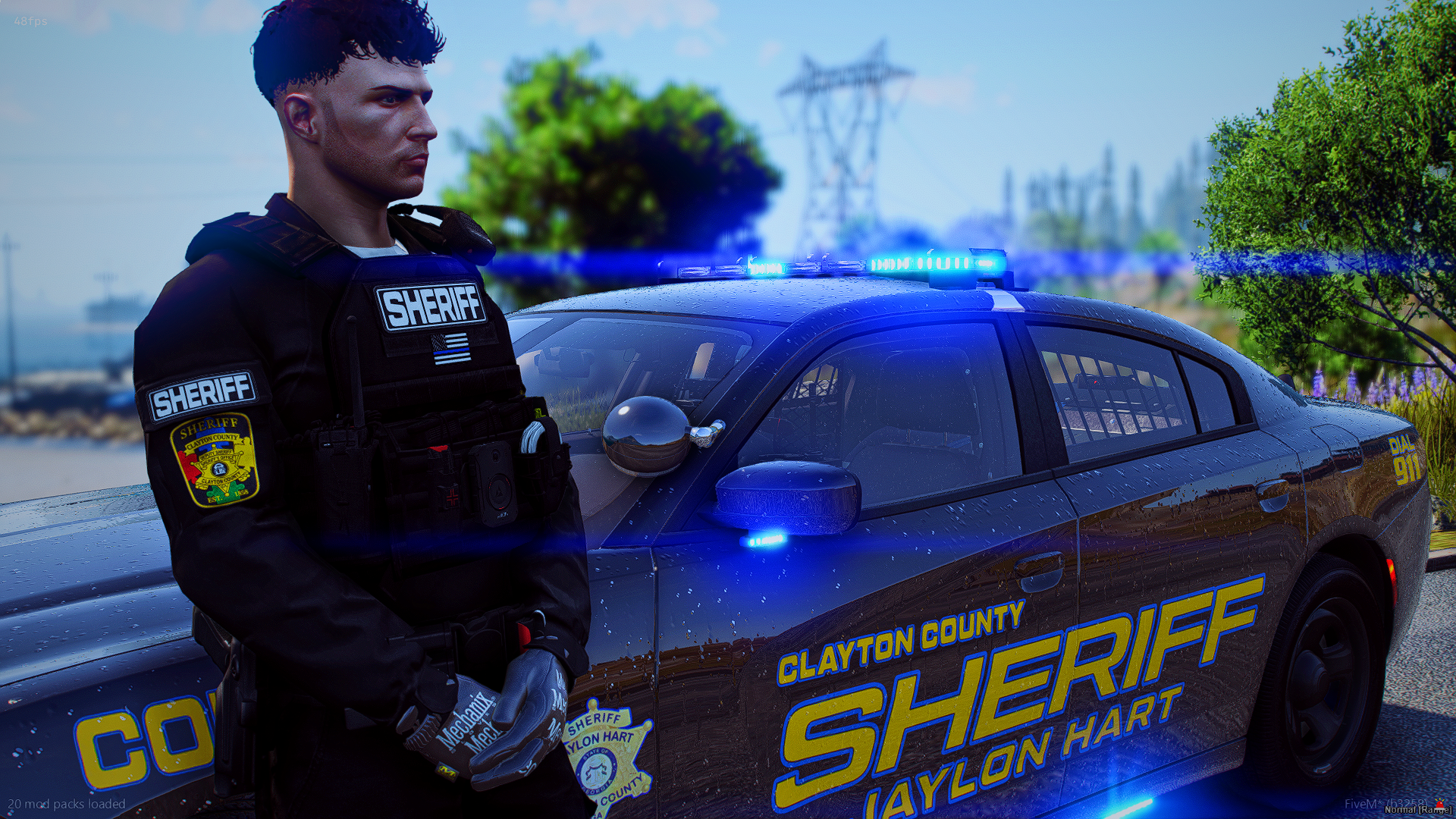 A police officer in a sheriff uniform standing beside a Clayton County sheriff patrol car with blue emergency lights on, outdoors near trees and power lines.