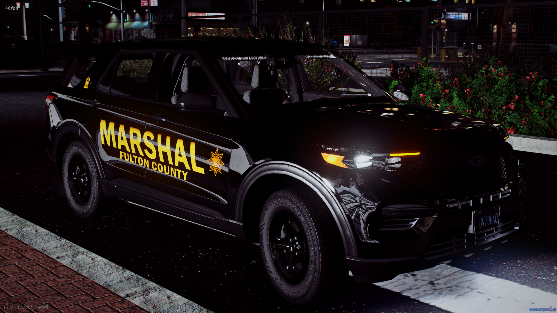 Nighttime image of a black sheriff's patrol vehicle with 'Marshal Fulton County' written on the side, parked on a city street with flowers and buildings in the background.