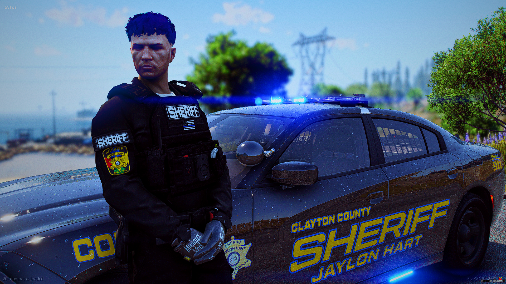 A police officer stands next to a Clayton County sheriff's vehicle with blue lights on, on a road near a body of water, with trees and power lines in the background.