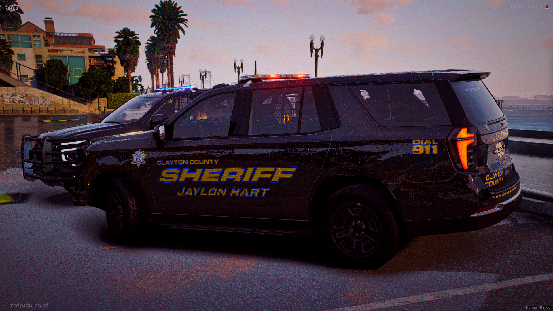 A black Clayton County Sheriff's vehicle with flashing red and blue lights parked near a waterfront with palm trees and colorful buildings in the background during sunset.