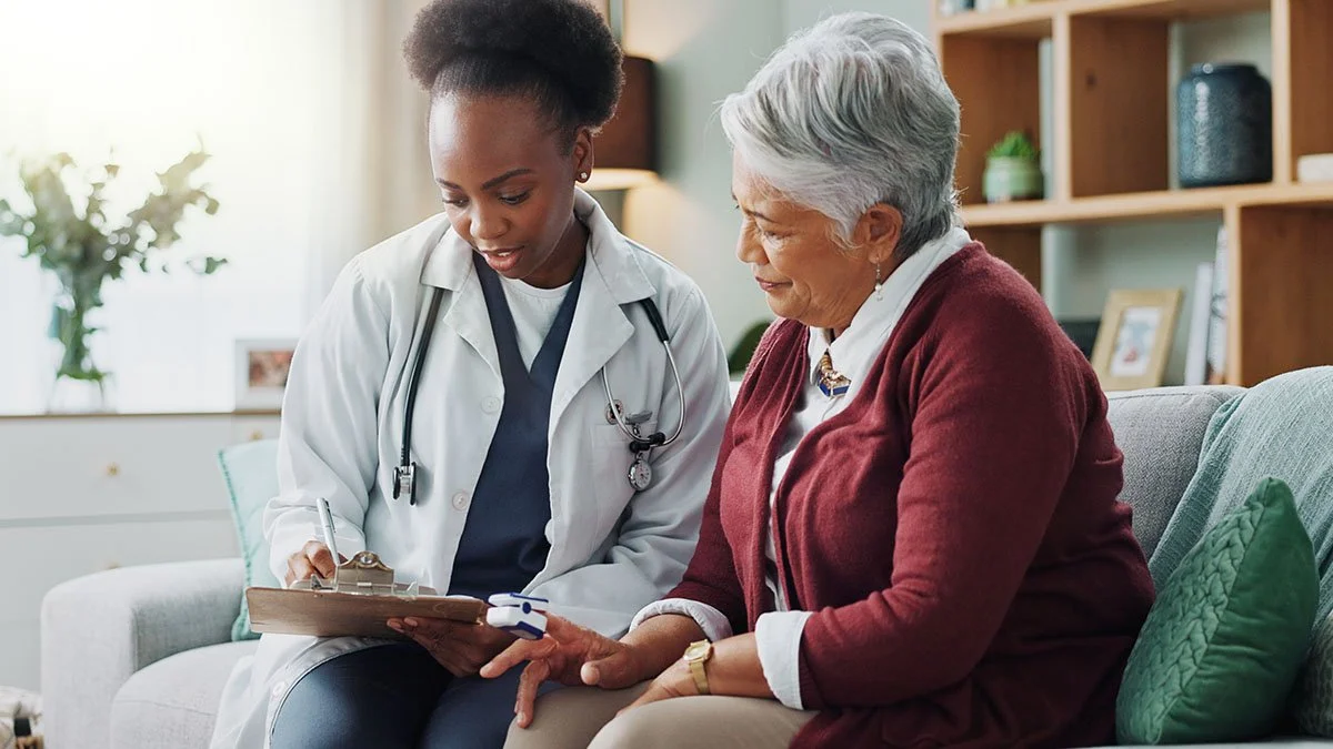 A female healthcare professional taking notes on a clipboard while talking to an elderly woman who is sitting on a couch, holding a blood pressure cuff in her hand in a living room.