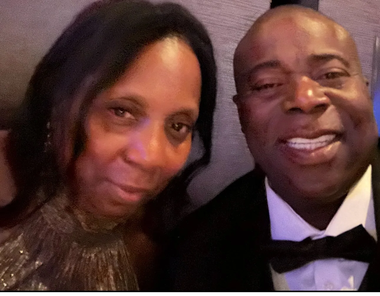 A man and woman in formal attire, smiling at the camera, sitting close together at a formal event.
