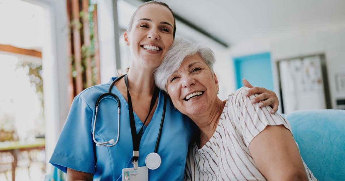 A young female nurse with a stethoscope around her neck smiling and hugging an elderly woman with short gray hair in a hospital or care facility.