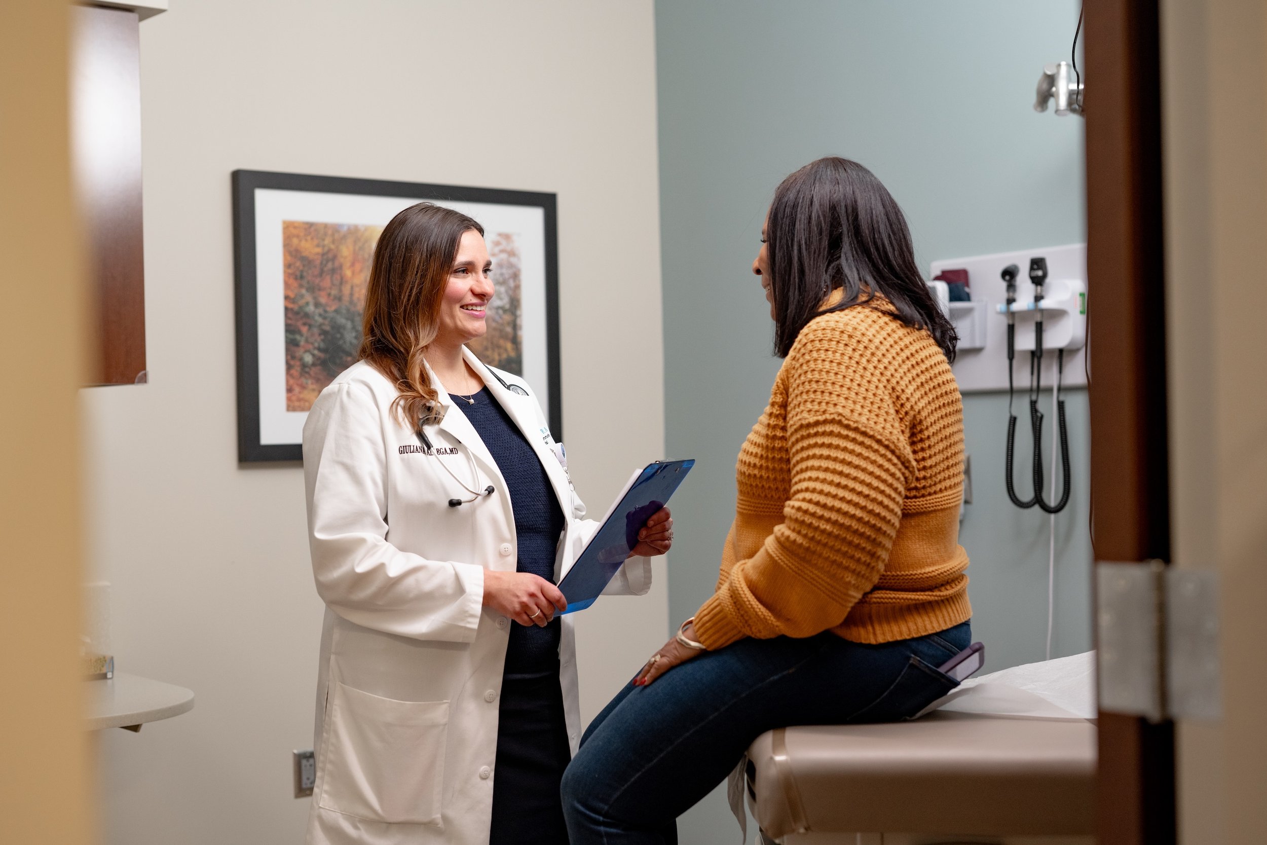 A female doctor speaking with a woman in a doctor's office, with a medical exam table and wall-mounted medical equipment in the background.