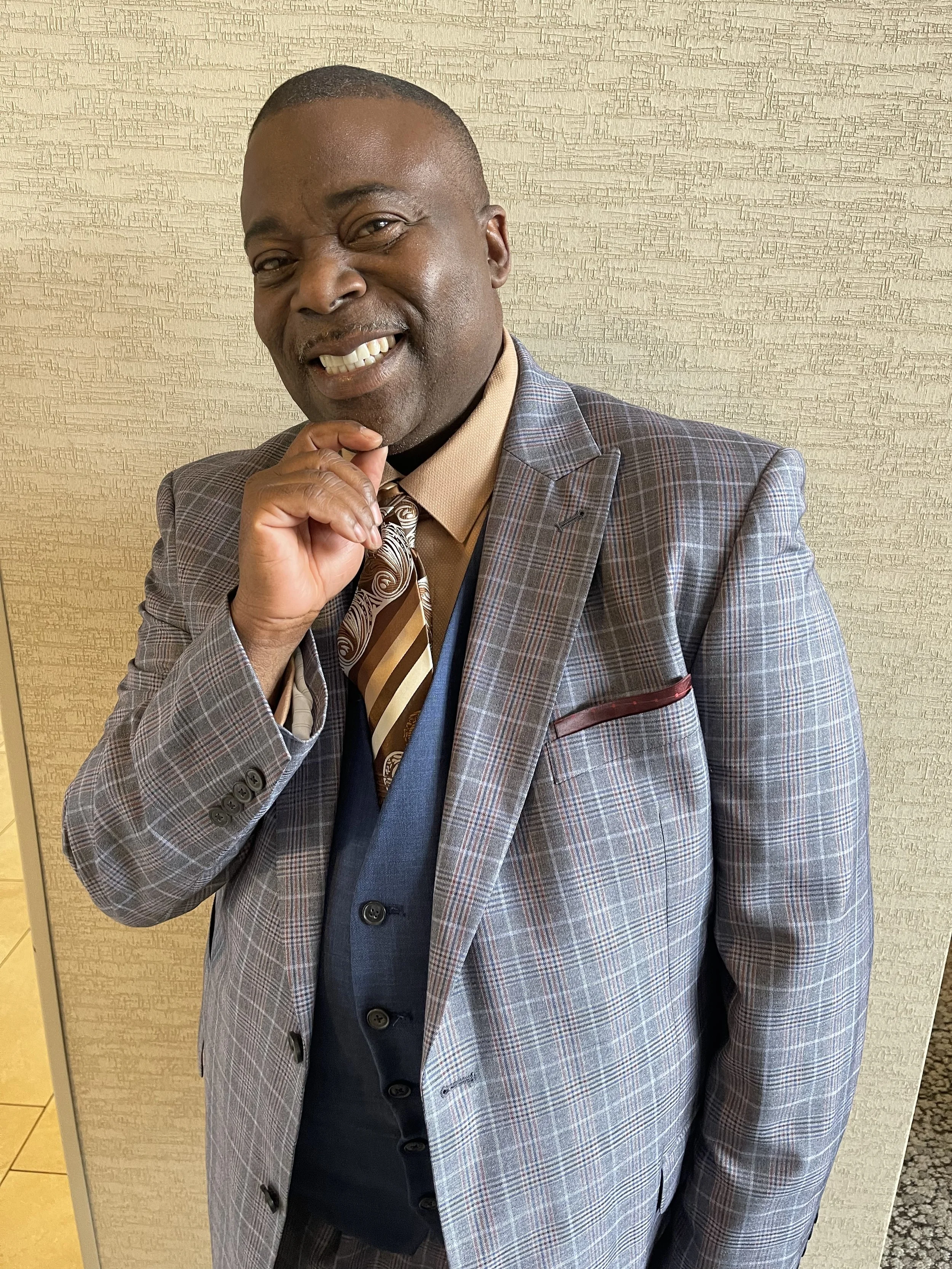 A man in a checked gray suit smiling and posing with his hand on his chin in front of a beige textured wall.