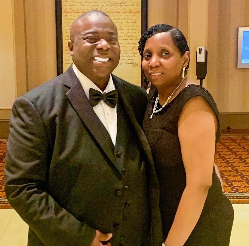 A man in a black tuxedo and bowtie and a woman in a black dress standing together indoors at a formal event.