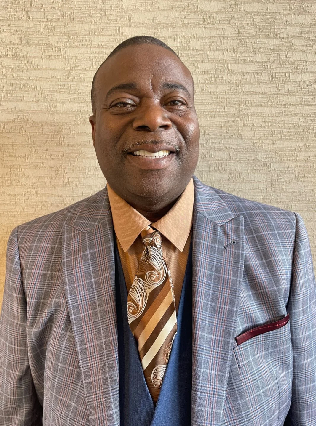 A smiling man in a plaid suit with a patterned tie and beige shirt, standing in front of a textured beige wall.