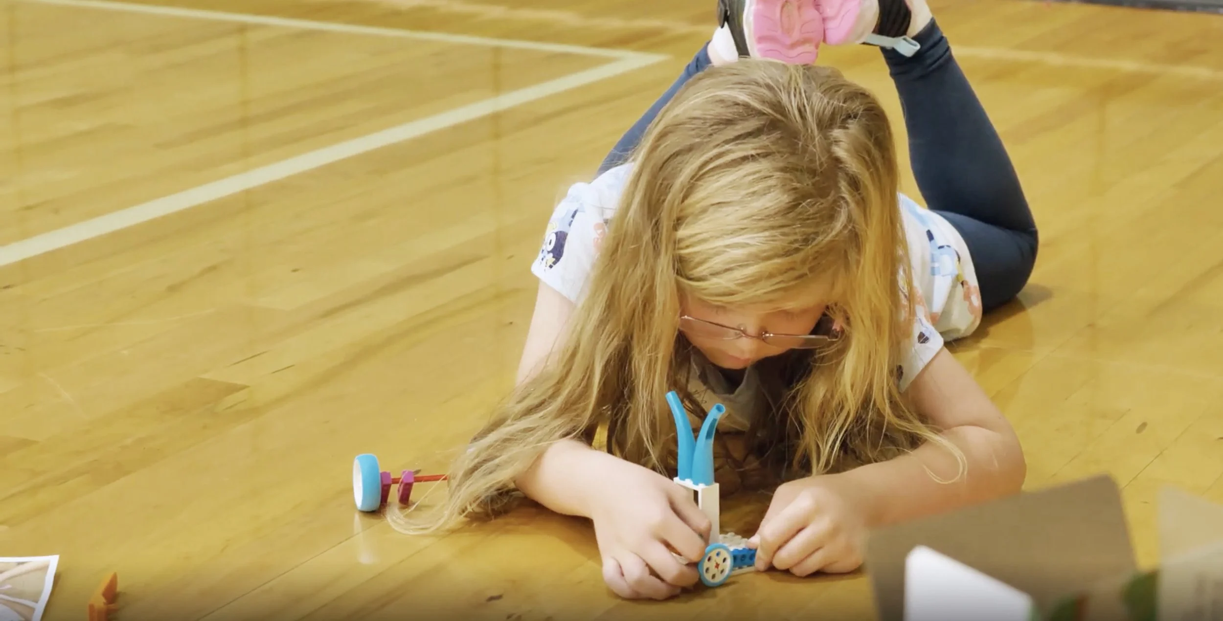 A young girl with long blonde hair, wearing glasses, is lying on a wooden gym floor, working on building a LEGO project, with a small LEGO vehicle beside her.