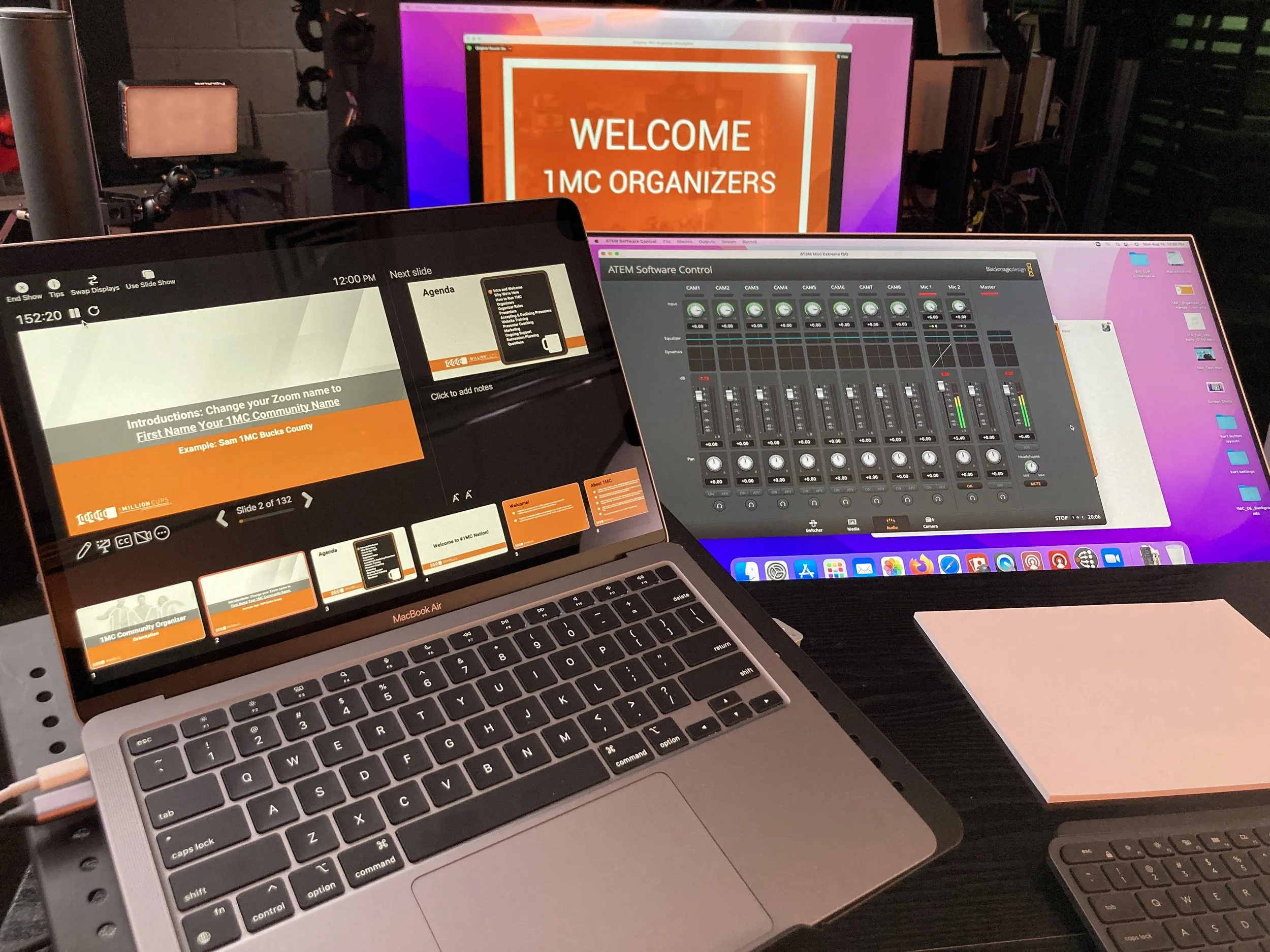 A laptop displaying a presentation slide with the title 'Introduction: Change your Zoom name to First Name Your 1MC Community Name' with an example name. In the background, there are two screens: one with a welcome message for 1MC organizers, and another displaying audio control settings.