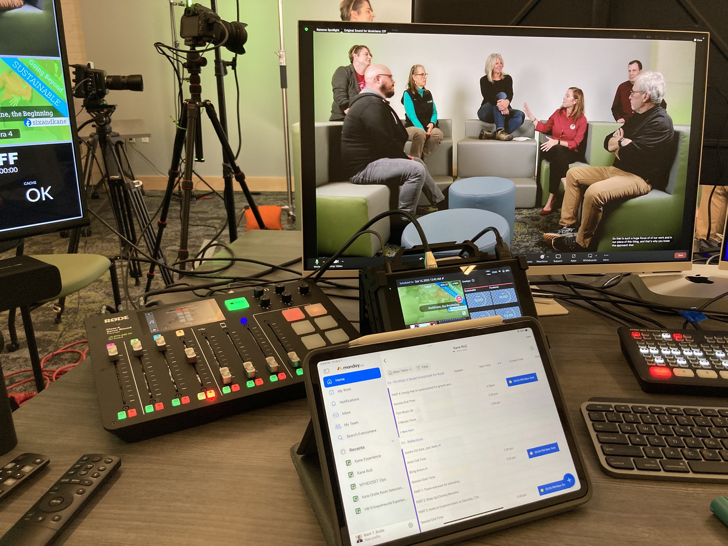 A video camera on tripod, studio lights, and various electronic equipment including a mixer, computer, and remote controls in a workspace. The workspace shows multiple screens and a panel with controls, indicating a video production or live streaming setup.