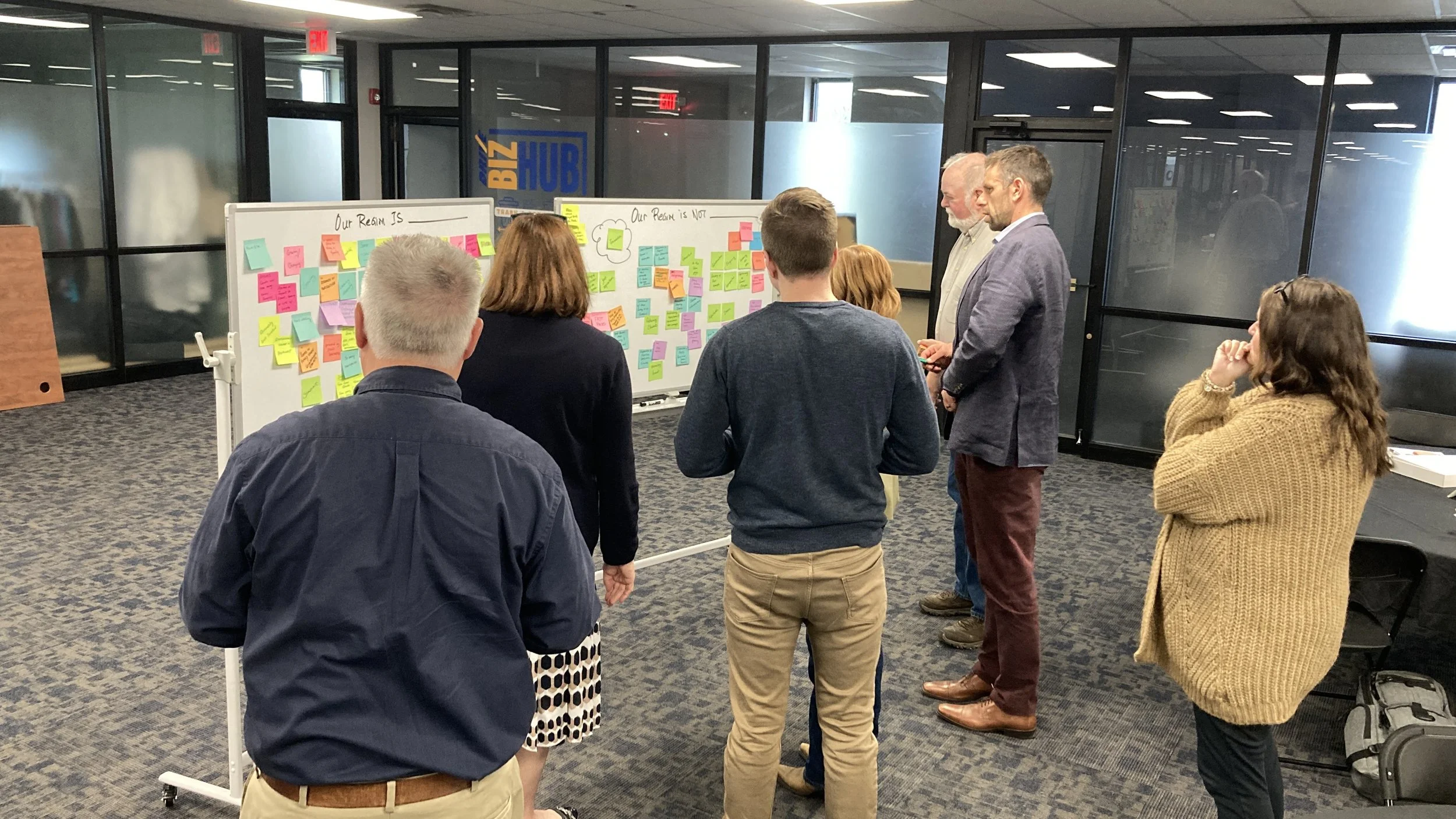 A group of people standing in a room looking at whiteboards covered with colorful sticky notes. The whiteboards have headings 'Our Vision IS' and 'Our Vision is NOT.' There are large glass windows and a glass door with the sign 'BIZ HUB' in the background.