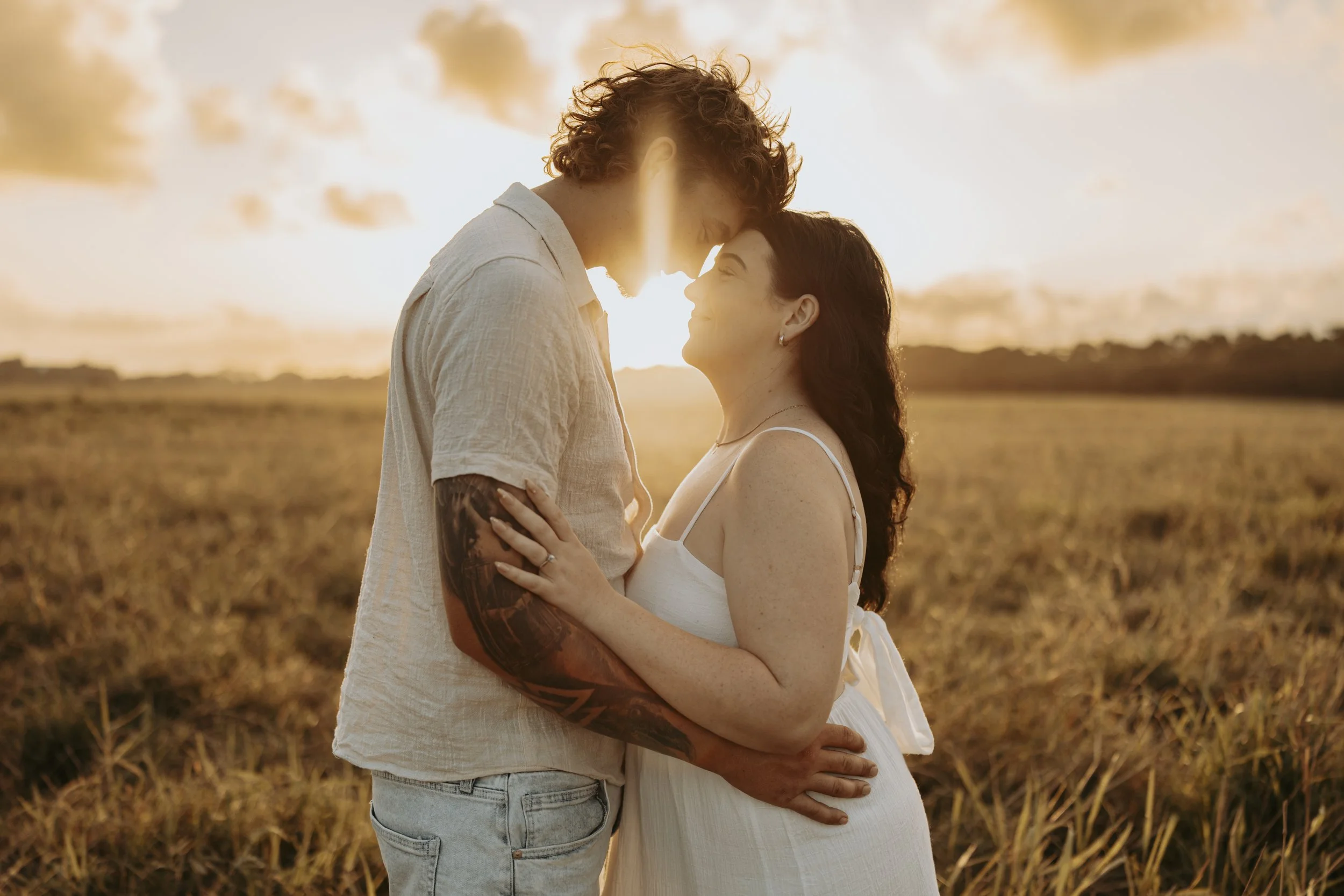 A couple standing close together in a field at sunset, facing each other with foreheads touching, holding each other gently, and smiling.