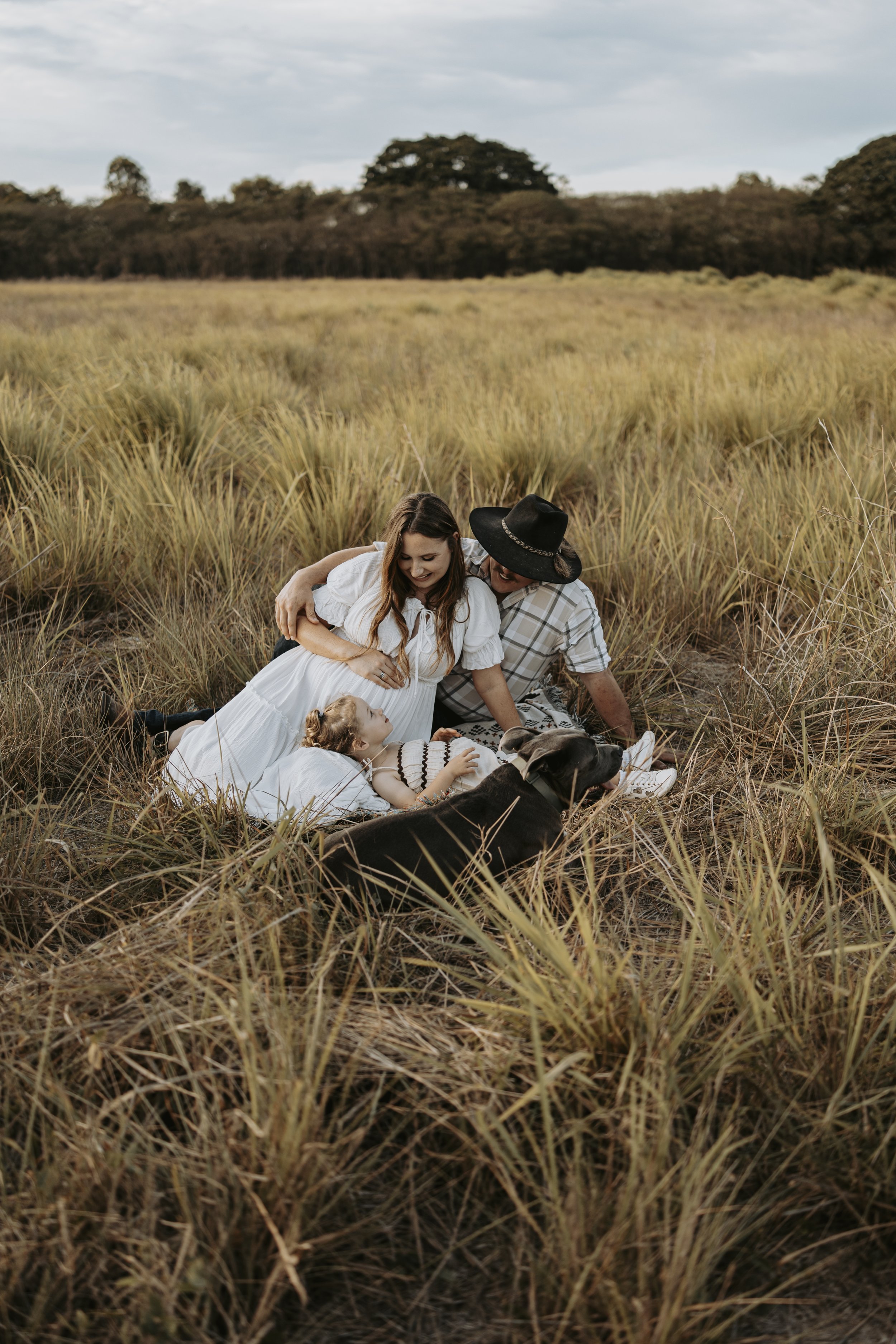 A family of four with a dog enjoying a picnic on a grassy field, with an open sky and trees in the background.
