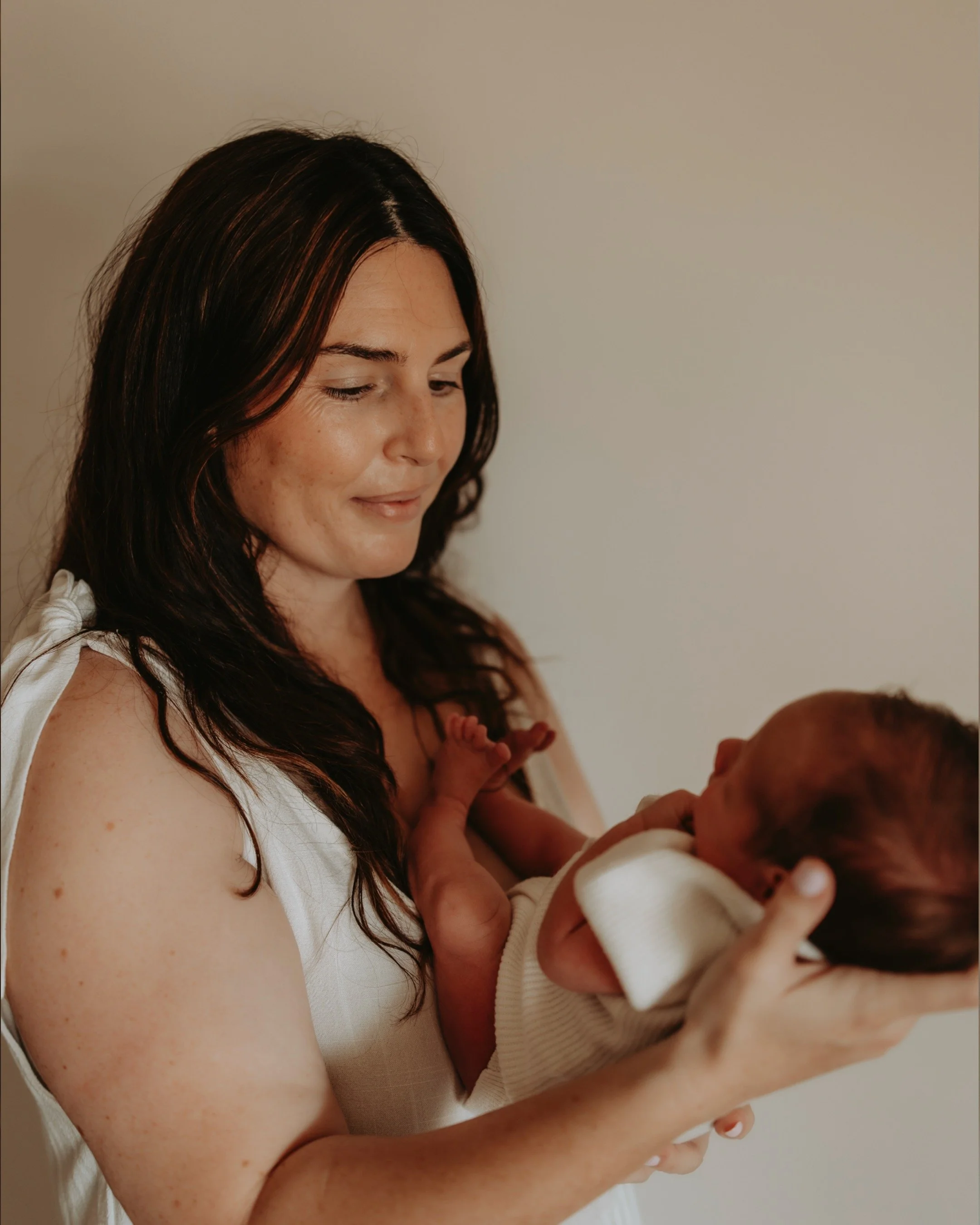 A woman with long dark hair holding a newborn baby with red hair. She is looking down at the baby, cradling the infant in her arms.