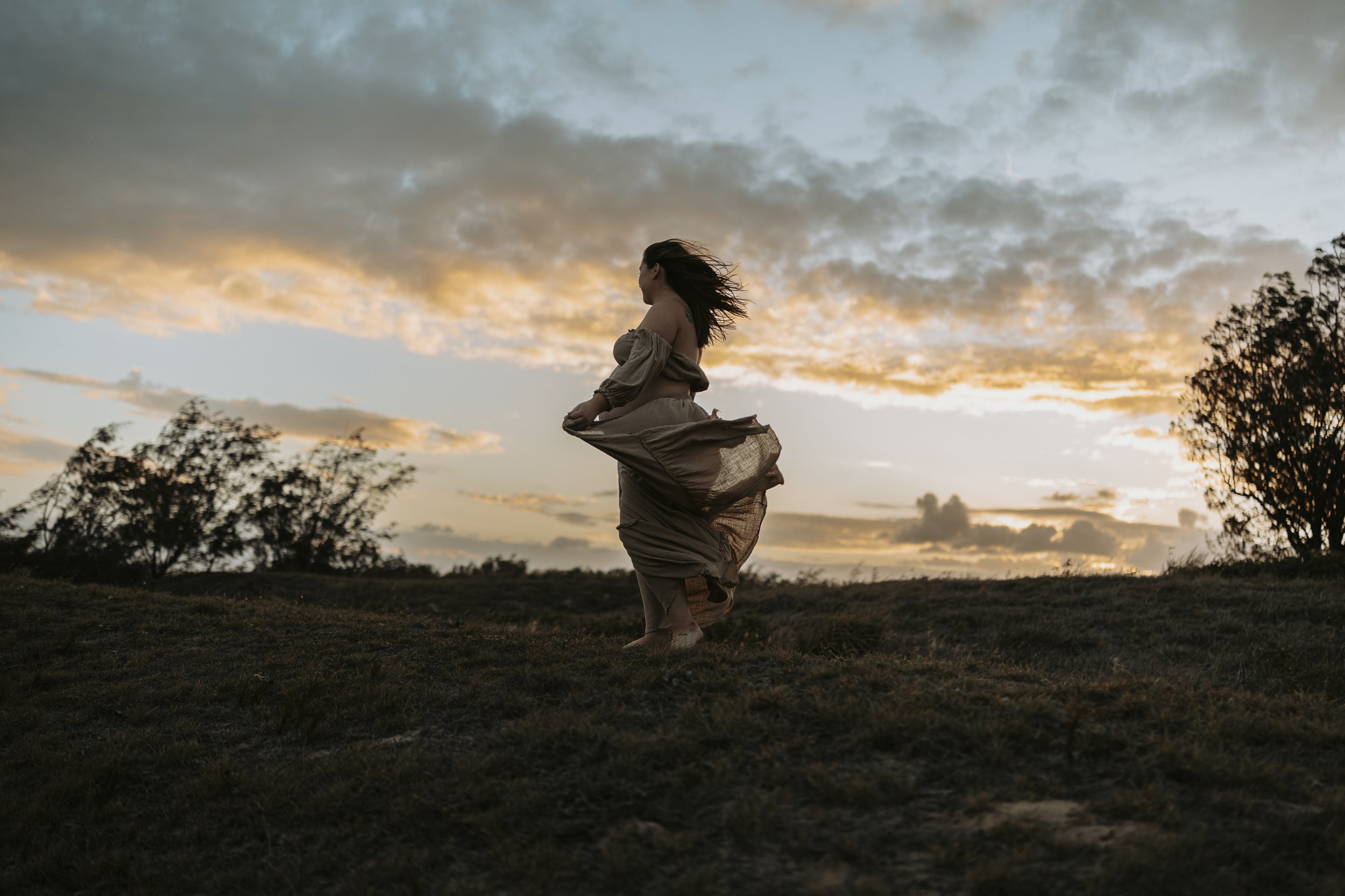 A woman in a flowing dress standing outdoors on a grassy hill during sunset, with a cloudy sky and trees in the background.