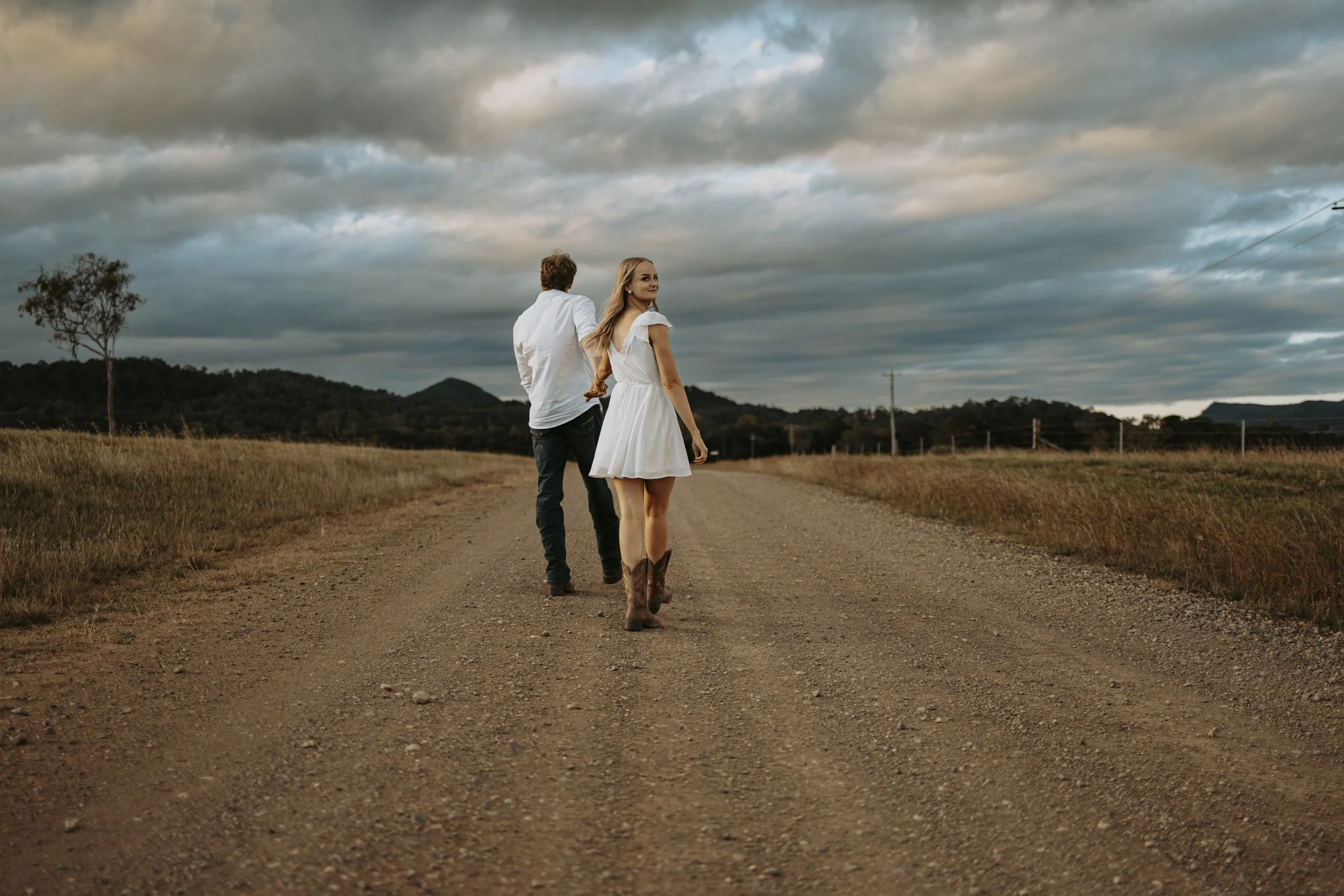 A woman in a white dress and cowboy boots holding hands with a man in a white shirt and dark jeans, walking on a dirt road through a field under a cloudy sky at sunset.