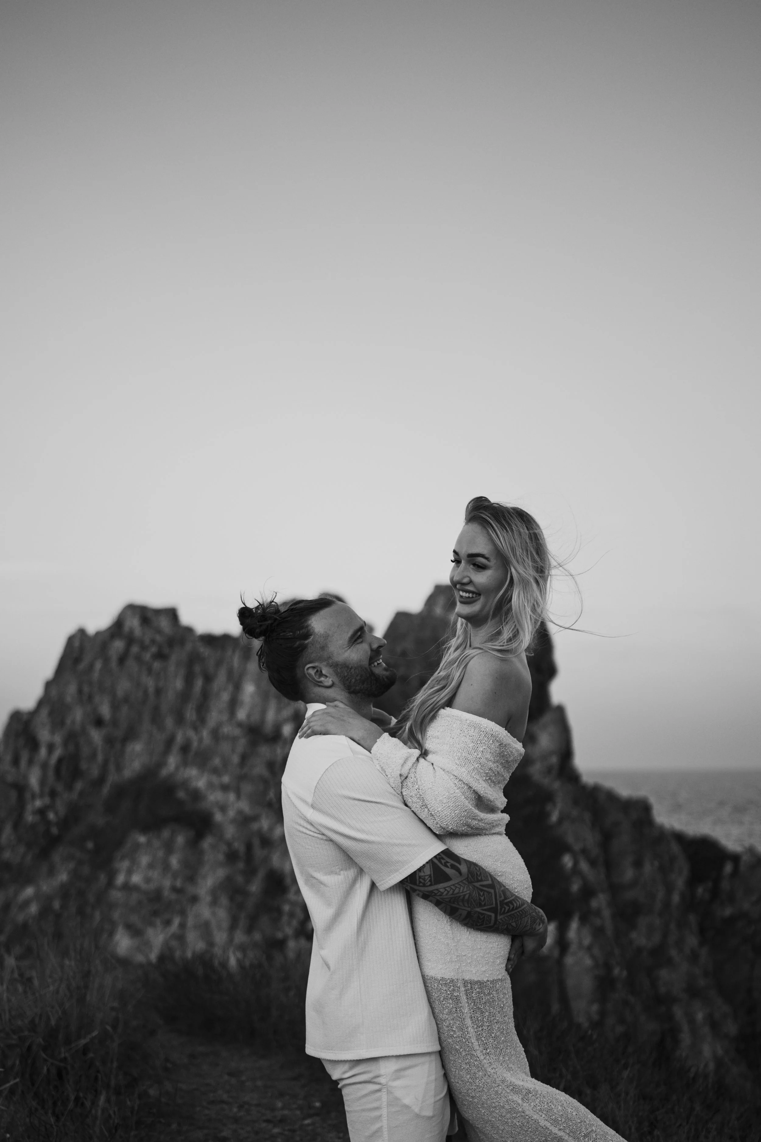 A black and white photo of a smiling couple outdoors near rocks and the ocean, with the man lifting the woman, both looking at each other.