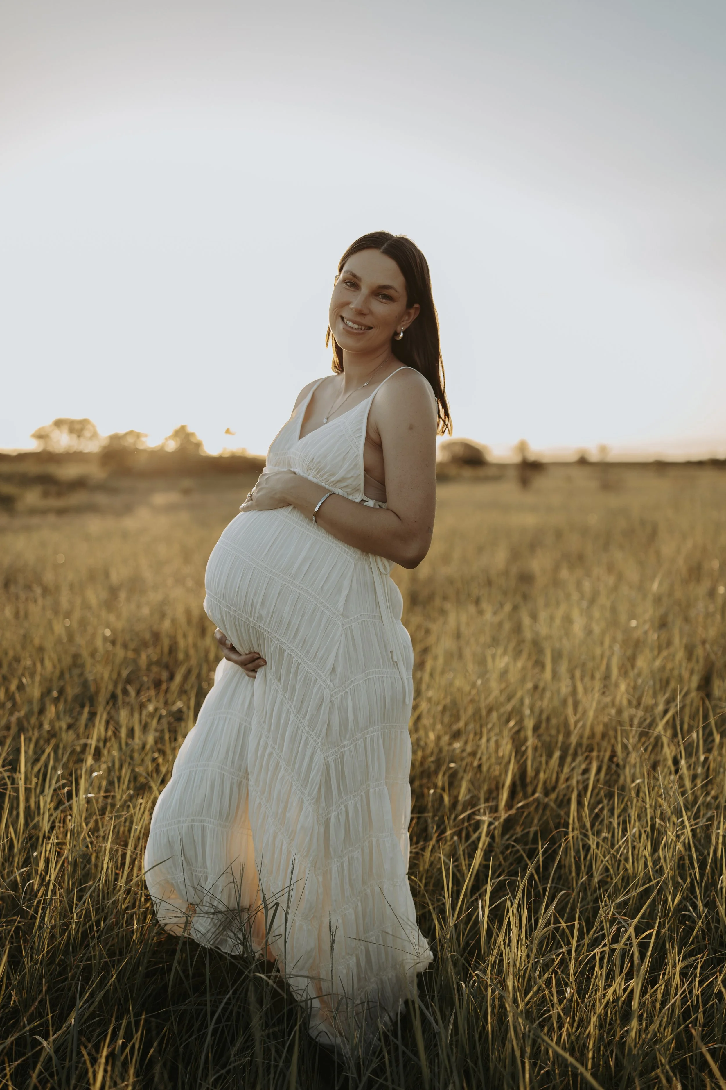 A pregnant woman in a white dress standing in a grassy field at sunset, smiling and holding her belly with one hand.