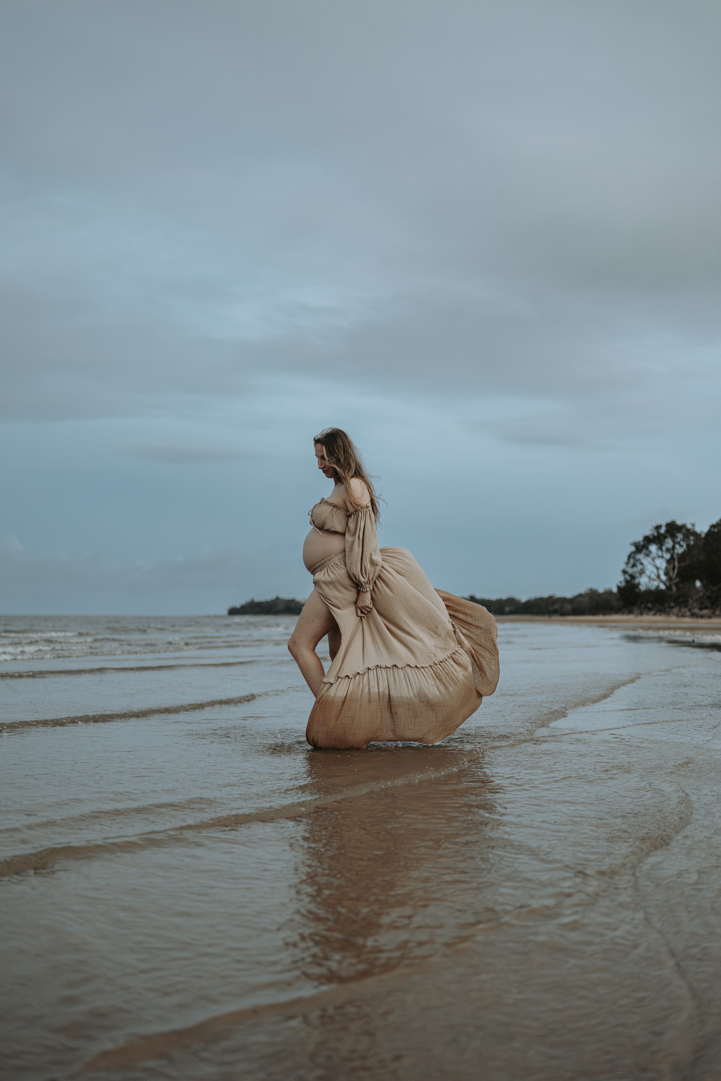 A pregnant woman wearing a flowing beige dress standing in shallow water at the beach during overcast weather.
