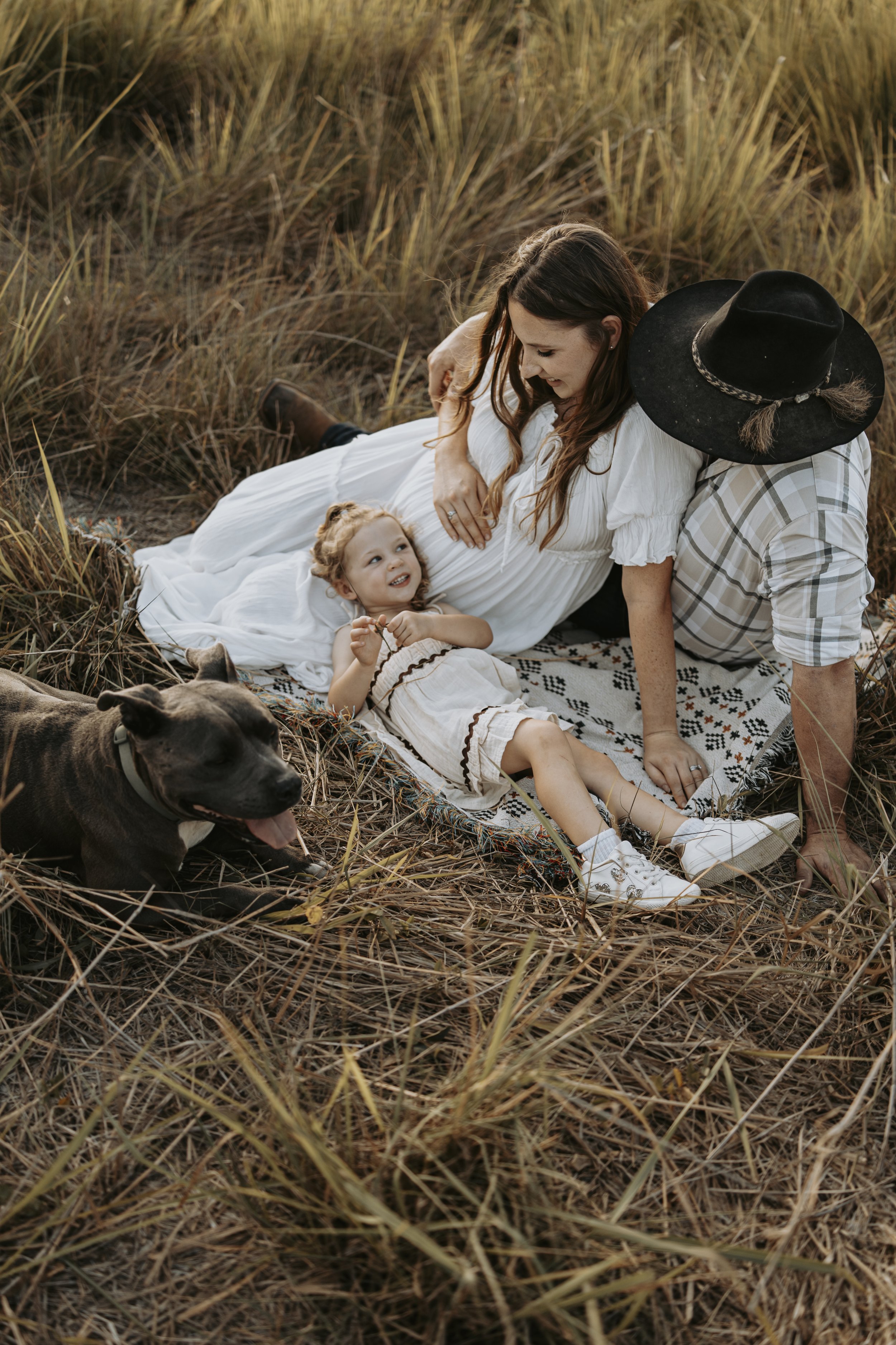 A family of three, including a mother, father, and young girl, lying on a patterned blanket in a field of tall grass during sunset. The mother, wearing a white dress, is smiling at the girl, who is lying down and looking up at her. The father, wearin