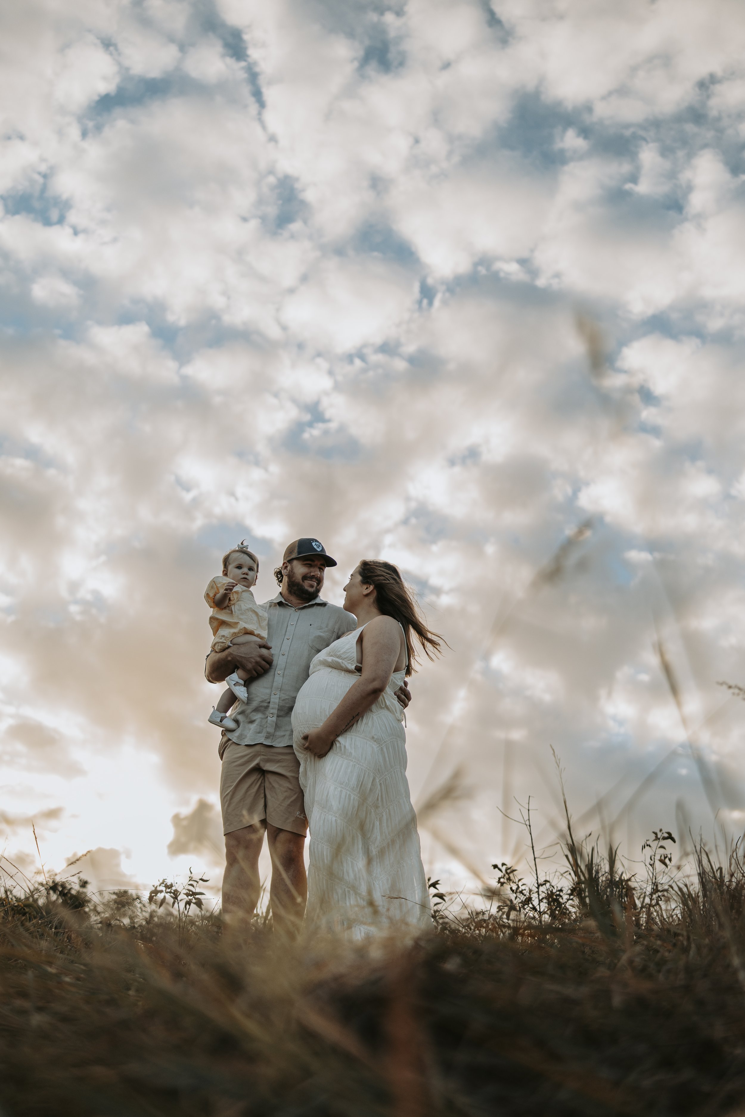 A family of three, a man, pregnant woman, and a small child, standing outdoors under a cloudy sky at sunset. The man is holding the child, who is pointing, and they are looking at each other and smiling.