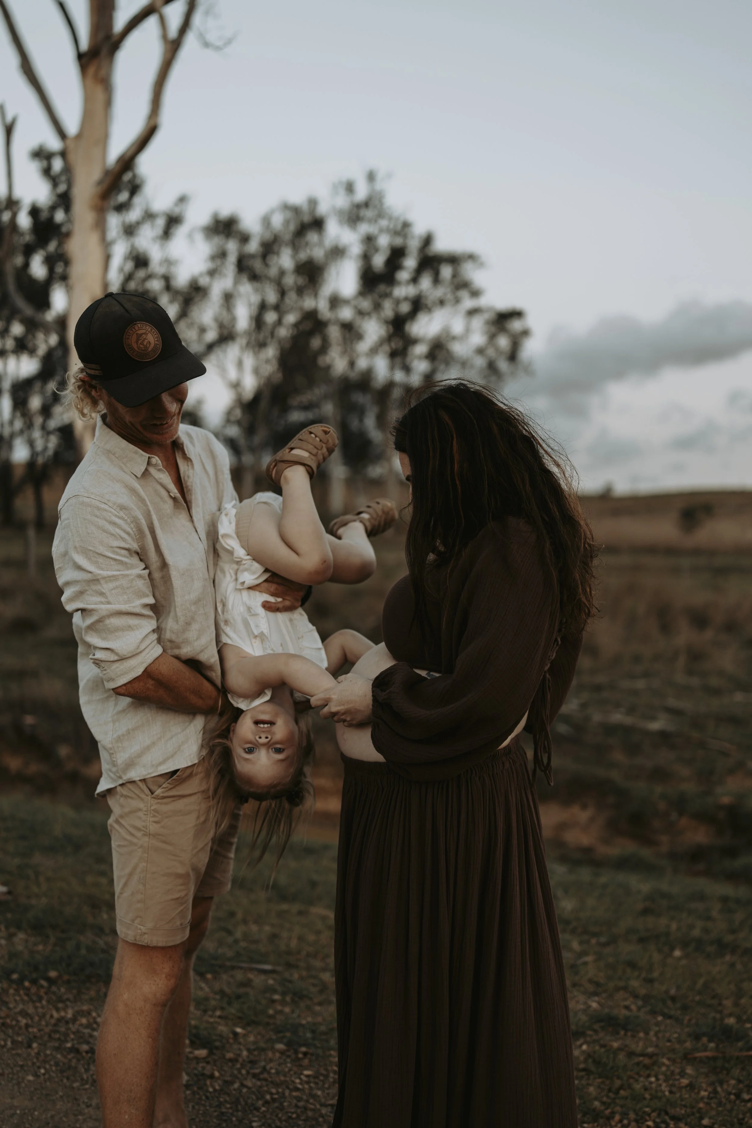 A man is holding a young girl upside down, while a woman stands nearby looking at the girl with a smile. The scene is outdoors during sunset in a rural area with trees and open fields in the background.